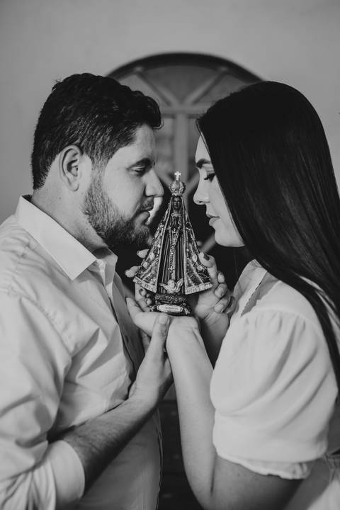 Fotografia em preto e branco de Jaque e Mateus diante de uma imagem religiosa durante o ensaio pré-casamento.'