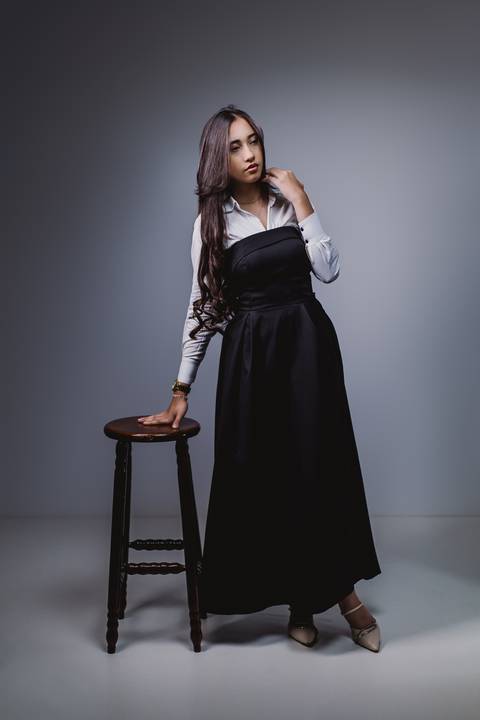 Retrato de corpo inteiro de Layla em estúdio, utilizando vestido preto e banco como elemento da composição.'
