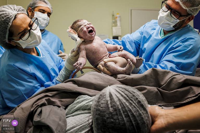 A Associação FPJA premiou Adriana Costa pela imagem que mostra a equipe médica num cuidado carinhoso de abaixar o pano durante a cesariana para que a mãe pudesse ver seu bebê assim que nascesse