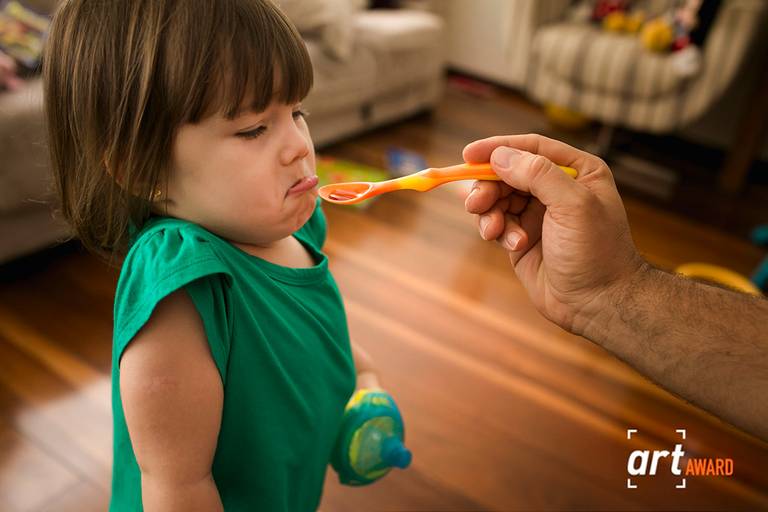 foto premiada de Adriana Costa pela Fineart Association de uma criança fazendo careta para o remédio dado pelo pai 