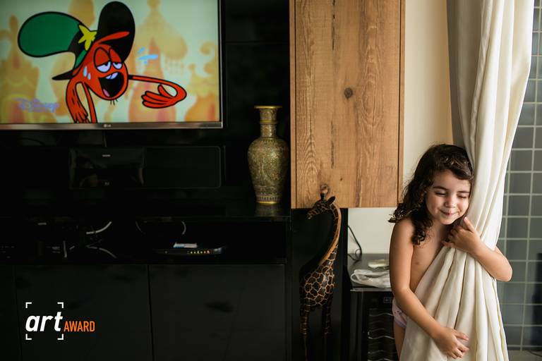 Foto de Adriana Costa premiada pela fineart association de uma menina abraçando a cortina e tendo ao fundo a imagem de um personagem na tv suando e com o mesmo gesto de abraçar da menina