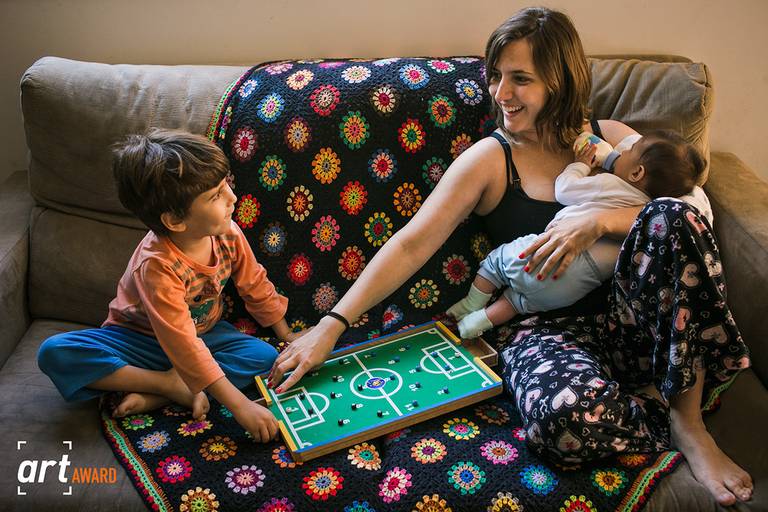foto premiada de Adriana Costa pela fineart association de uma mãe amamentando seu bebê ao mesmo tempo que entretêm seu filho mais velho com um jogo de futebol de botão