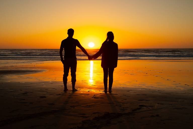 namorados de mão dada na praia a observar o por do sol