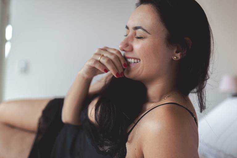 mulher deitada na cama, de camisola preta, sorrindo com a mão na boca