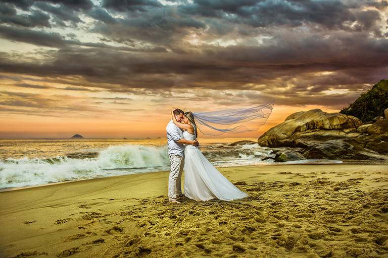 noivos na areia da praia com o céu nublado no fim de tarde, noiva com o véu voando e ondas ao fundo durante o ensaio trash the dress, Niterói, Rio de Janeiro, RJ