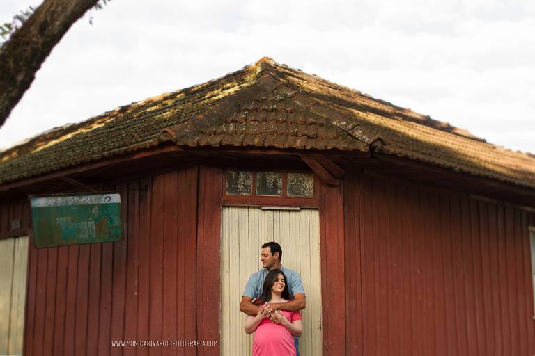 Em frente a uma casa de madeira antiga onde funciona uma venda, o casal esta abraçado, ele atrás dela e olhando para a esquerda e ela na frente e olhando para direita. Nesta venda, a gestante comprava doces quando era pequena