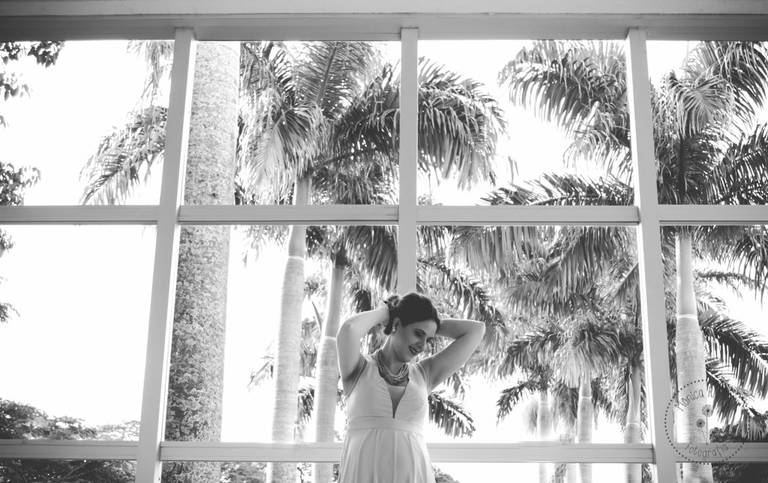 Gestante prendendo seu cabelo e sorrindo, ao fundo uma parde de vidro e atrás coqueiros