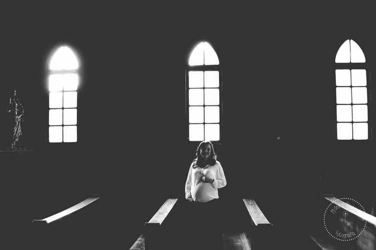 Gestante no meio de uma capela, posando com as mãos no seu bebê, foto preto e branco