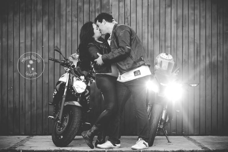 Casal se beijando no meio de duas motos, ao fundo uma parede de madeira, foto preto e branco