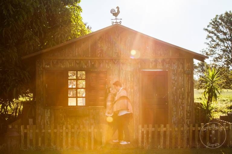Casal se abraçando, envoltos por uma manta, ao fundo uma casinha de madeira, brilha um sol lindo atravessando a casinha