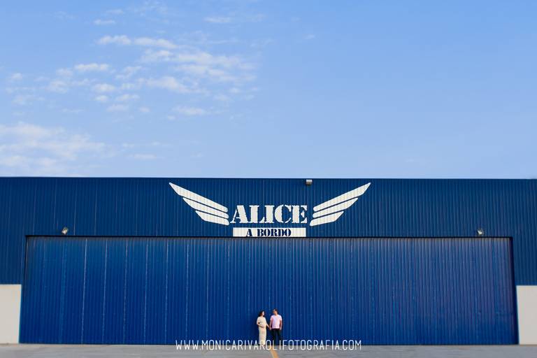 Casal se olhando em frente a um grande galpão azul, com o nome da sua bebê em cima