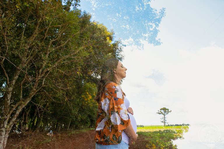 A Nayara olha para cima segurando seu bebê, a foto foi feita com efeito de dupla exposição, onde o corpo da Nayara faz contorno das árvores atrás dela
