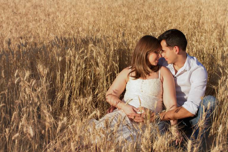 Casal sentando no trigo, com as mãos no bebê e com os rosto encostado um no outro.  
