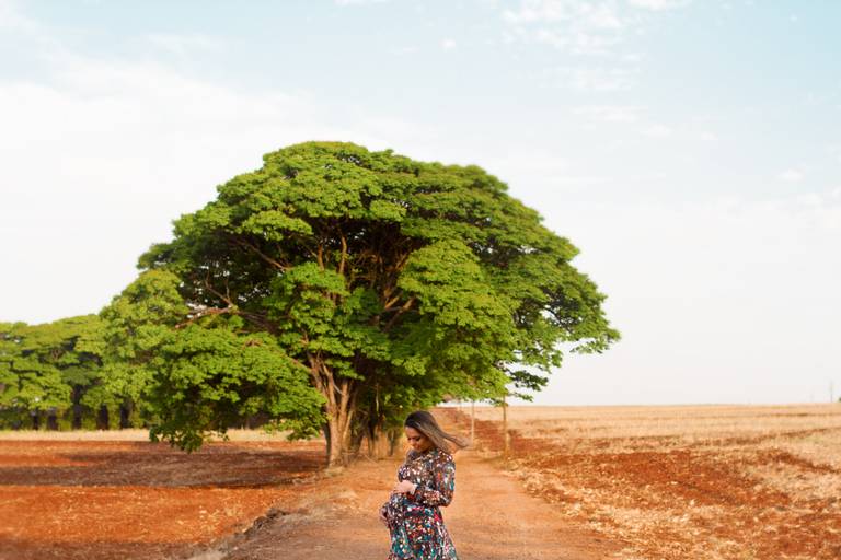 Nesta fotografia, a gestante está em uma fazenda nas proximidades de Maringá/PR. Ao fundo, há uma árvore de copa larga e alta com muitas folhas verdes. A gestante apoia as mãos na barriga e, olhando para seu bebê, ela sorri