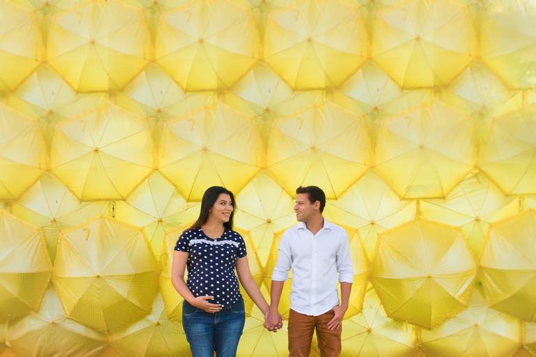 Nesta fotografia, o casal está de de mãos dadas se olhando, A gestante com a mão na barriga. Ao fundo uma parede de guarda-chuvas amarelo