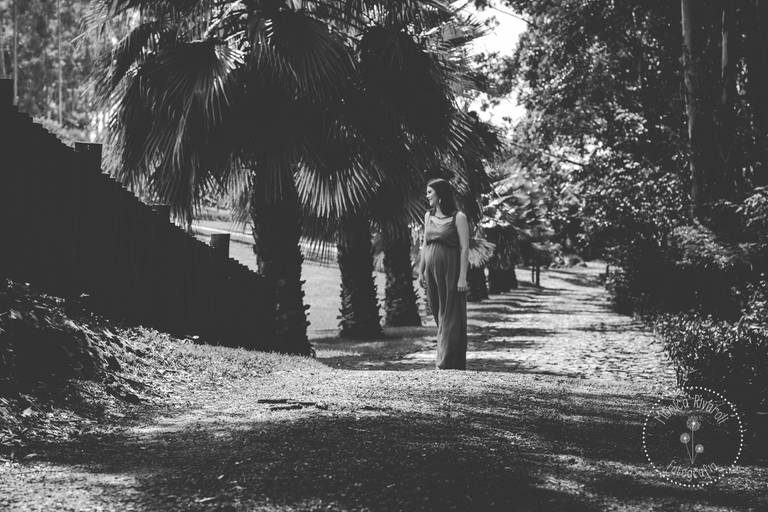 Na foto, gestante ao centro caminhando por um corredor cheio de arvores, com um sorriso no rosto. No canto direito inferior a logo da fotografa Mônica Rivaroli. Foto preto e branco 