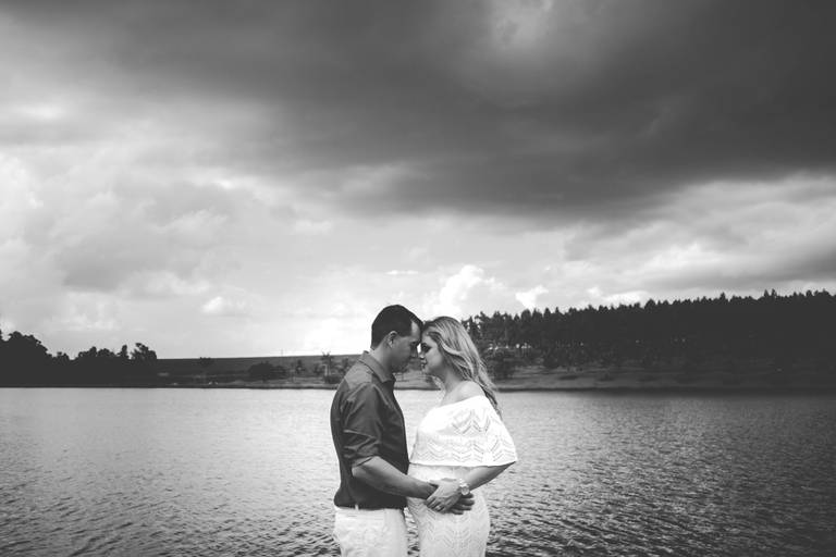 A imagem mostra o perfil do casal, com as testas encostadas, o marido com as mãos no bebê dentro da barriga da mamãe. Ao fundo um lago e árvores no horizonte. Foto preto e branco.  