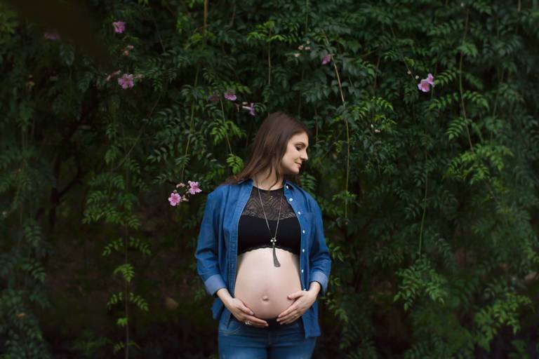 Gestante com um top preto, mostrando sua barriga, com as mãos no seu bebê, ao fundo uma parede com folhas verdes e flores 