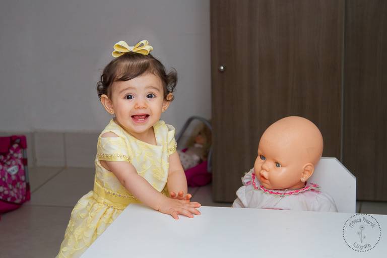Isabelle sorrindo , com um lindo vestido amarelo e um lacinho da mesma cor no cabelinho, ao lado uma boneca sentada na cadeirinha e ela com as mãozinhas na mesa 
