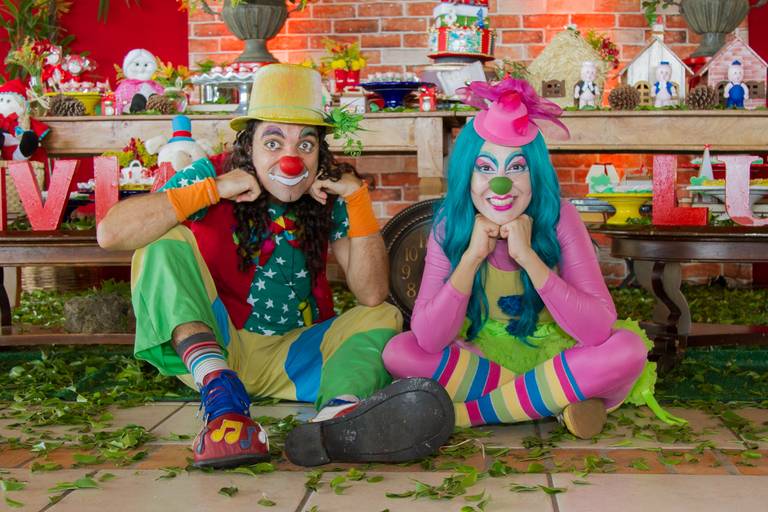 Piruca de Vagalume sentados em frente a decoração da festinha da Lívia e do Lucas. Pipoca fazendo careta e Spirulina sentada de indinho, com as mãos no queixo sorrindo 