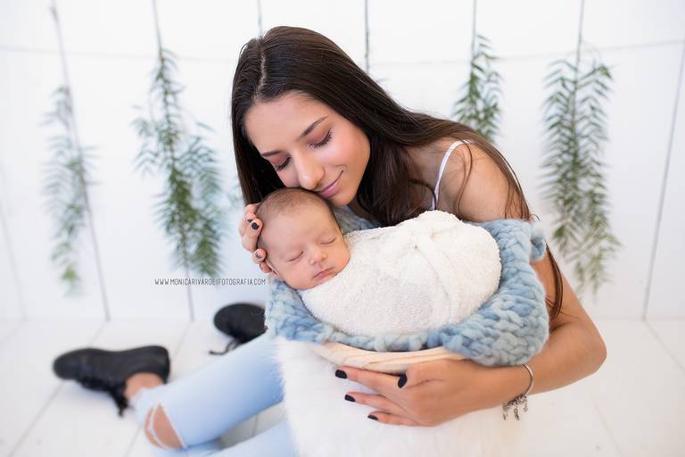 Nesta fotografia, estão o bebê e sua irmã mais velha. Ela o segura deitadinho em uma gamela que está posicionada em um mini banco de plumas fofinhas. A irmã tem 15 anos de idade e está com o rosto próximo ao do irmão