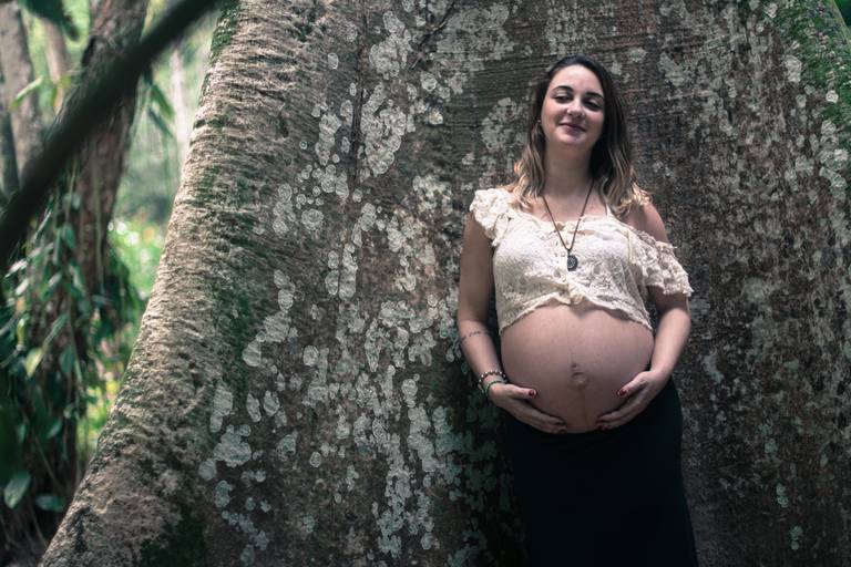 Grávida segurando a barriga de olhos fechados com um sorriso no rosto apiada em um tronco de árvore no Jardim Botânico no Rio de Janeiro