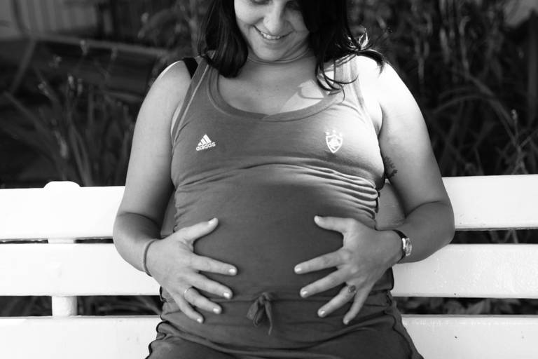 Gestante segurando a barriga e olhando para ela sentada em um banco no Fluminense Football Club no Rio de Janeiro