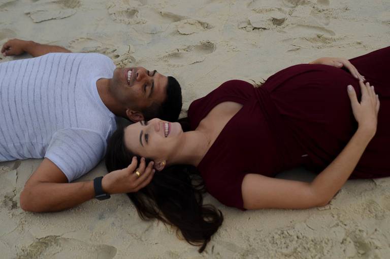 Casal Grávido sorrindo  deitados na areia no Quebra Mar na Praia da Barra da Tijuca no Rio de Janeiro
