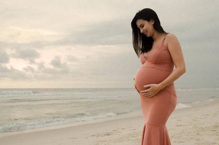 Gestante sorrindo segurando a barriga em frente ao mar  no Quebra Mar na Praia da Barra da Tijuca no Rio de Janeiro