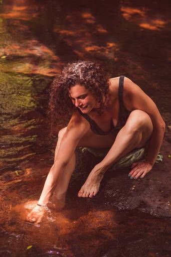 Modelo sentada em uma pedra no meio de um riacho tocando a água em um spot de luz 