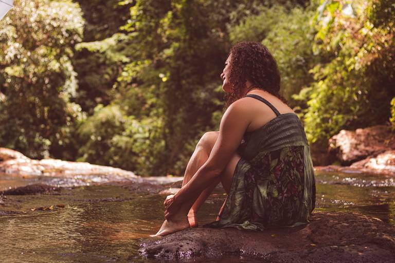 Modelo sentada em uma pedra no meio de um riacho de costas para câmera olhando para frente com os braços abraçando as pernas 