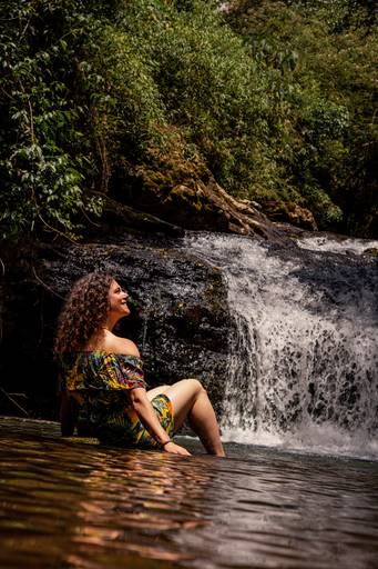 Mulher com vestido florido tomara que caia sentada no riacho e de frete para uma queda de cachoeira pequena