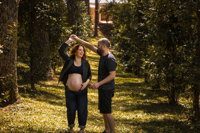 Gestante vestindo blazer e top preto acompanhada do marido vestindo roupas escuras girando ela em um passo de dança