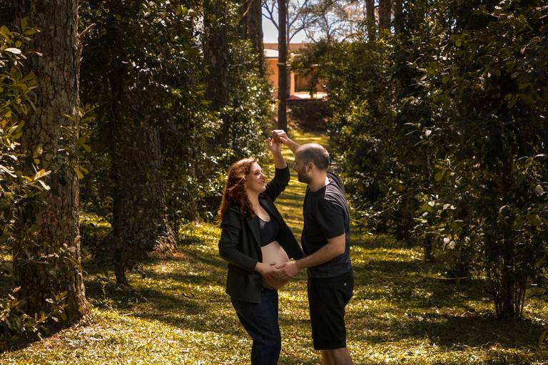 Gestante vestindo blazer e top preto acompanhada do marido vestindo roupas escuras dançando um de frente para o outro