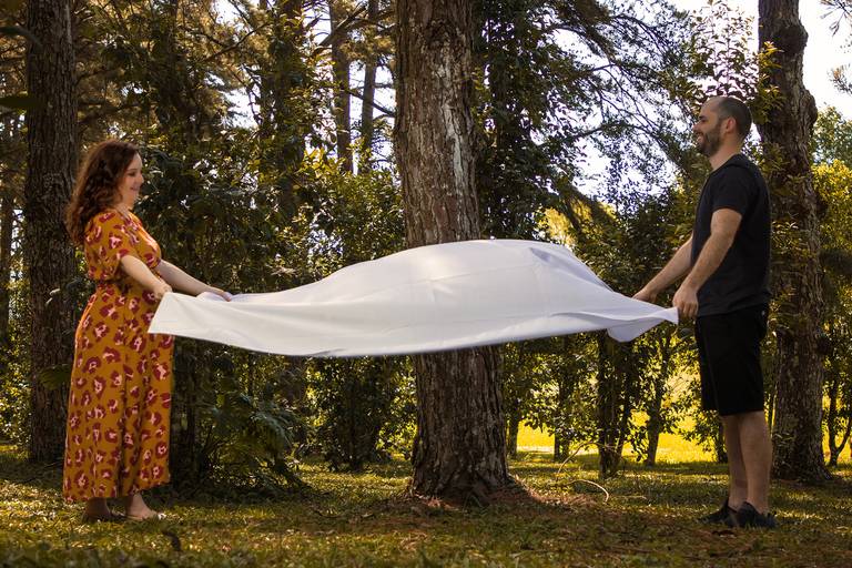 Mulher gestante usando vestido alaranjado florido e marido com roupas escuras estendendo pano branco no gramado para piquenique