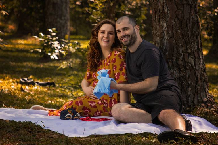 Mulher gestante usando vestido alaranjado florido e marido com roupas escuras sentados em cima de pano branco À sombra de uma árvore em um gramado