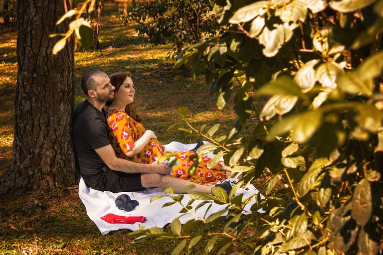 Mulher gestante usando vestido alaranjado florido sentada com as costas apoiada no marido com roupas escuras escorado em árvore sob um tecido branco no gramado