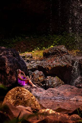 Mulher trajando vestido lilás sentada à sombra de uma pedra olhando a queda de uma cachoeira
