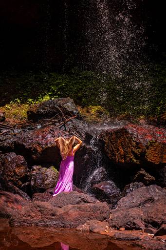Mulher trajando vestido lilás de pé em frente à uma queda de cachoeira mexendo no cabelo deixando em formato de coração