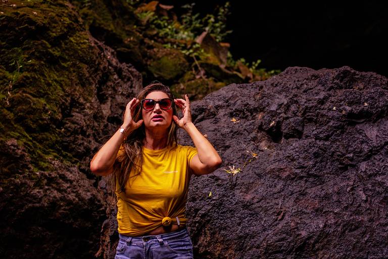 Modelo vestindo blusa amarela e short jeans colocando óculos escuros entre as pedras de uma cachoeira com pose de patroa