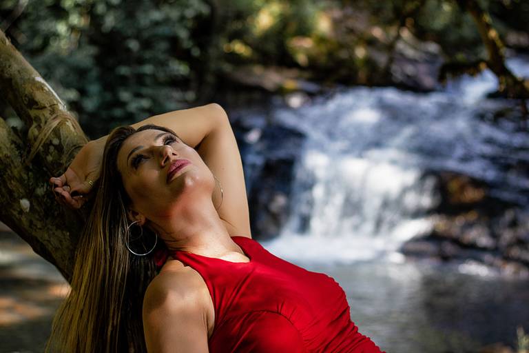 Mulher usando macacão vermelho deitada em um tronco de árvore com queda d'água ao fundo