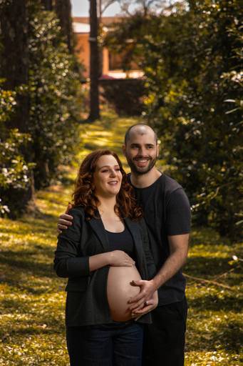 Gestante vestindo blazer e top preto acompanhada do marido vestindo roupas escuras se abraçando em área verde e olhando para o horizonte
