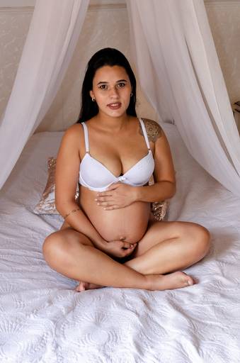 Ensaio de gestante da Veronica com lingerie branca, sentada na cama.
