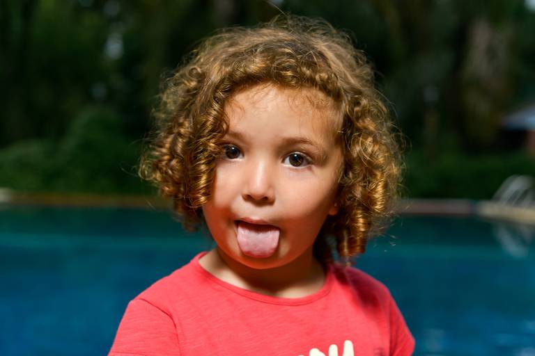 Niña con rulos sacando la lengua. Retrato infantil en exterior día de lluvia con una piscina de fondo. Marcos Pasquare fotografía de