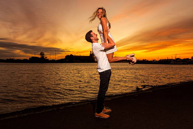 El novio la levanta a ella que sonríe para la cámara con un hermoso amanecer. Sesión pre boda. Marcos Pasquare Fotógrafo de Bodas.