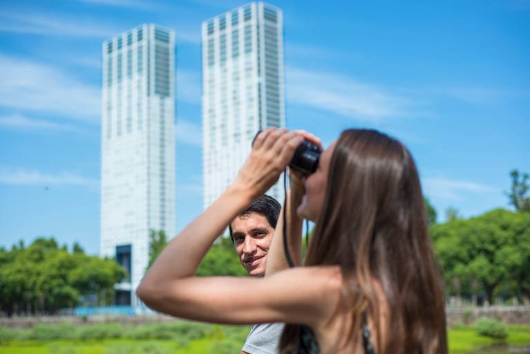Novia mira por binoculares y el novio la mira a ella. Sesión pre boda en la Costanera Sur, CABA. Reserva ecológica. Marcos Pasquare Fotógrafo de Bodas.