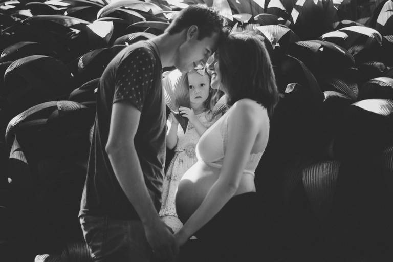 Foto em preto e branco com casal de testas e mãos unidas e criança no meio deles ao fundo
