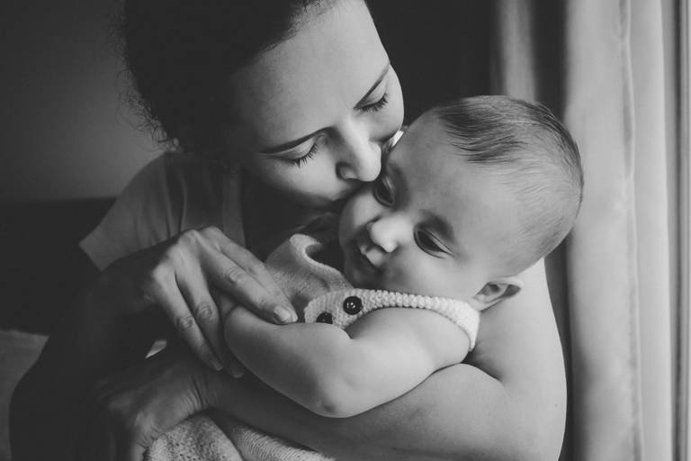 foto em preto e branco de mãe beijando bebe ao lado da janela em casa