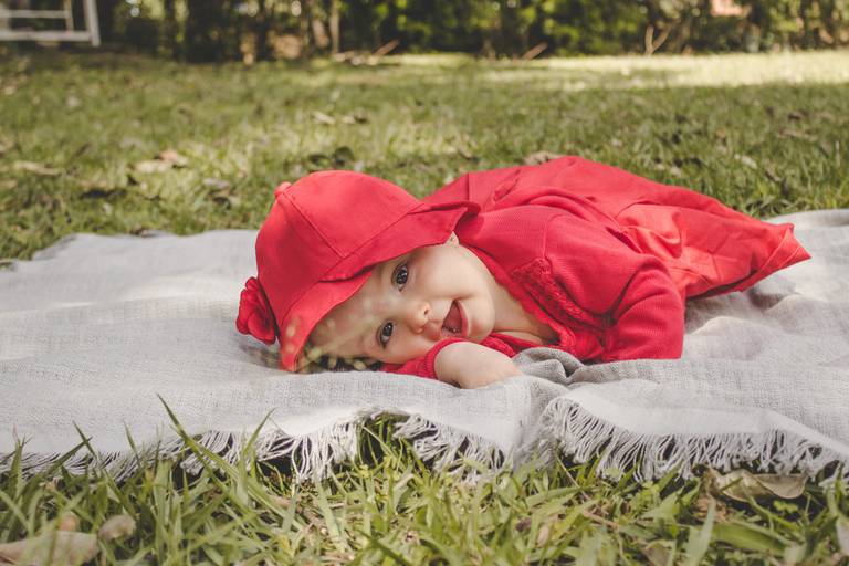 Bebê deitada no gramado sobre manta fazendo pose fofa  de chapeuzinho vermelho