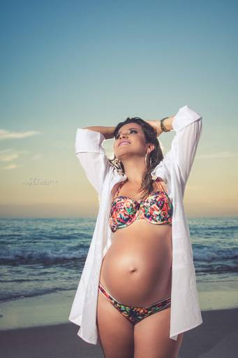 Roberta Lopes em posando sorridente para ensaio gestante de bikini estampado e blusa branca na Praia de Grumari.